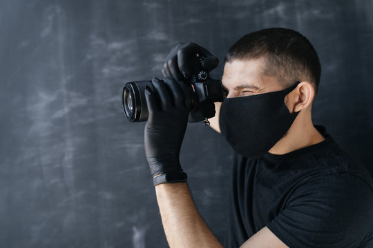 man Photographer in a protective black mask and gloves holds a camera photographs on a dark background. Photo business, coronovirus.