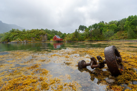 Rusty winch on a seaweed-covered shore near Hadsel, Norway, with calm water, green hills and cloudy sky.