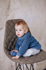 Fototapeta premium Cute Studio portrait of a one-year-old boy sitting on a chair. A boy in a blue sweater and beige pants