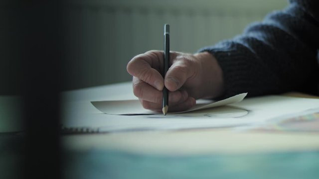 A 4K shot of a mans hand drawing a picture with a pencil. Shot from the side with shallow depth of field of an older man sketching. Wider shot of hand.