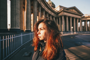 A young brunette girl with long hair in a jacket stands in the rays of the morning dawn on a...