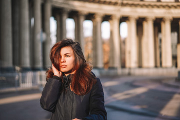 Fototapeta premium A young brunette girl with long hair in a jacket stands in the rays of the morning dawn on a deserted square in St. Petersburg against the background of the columns of the Kazan Cathedral