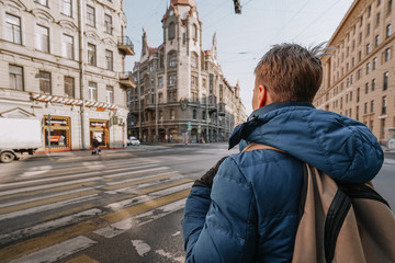 A young man in a jacket and a backpack stands at a traffic light against the background of an architectural monument of Federal significance, an administrative building located in the center of St. Pe