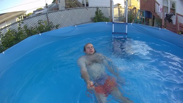 Slow motion of man in orange shorts recording hims self while swimming on his back in back yard swimming pool