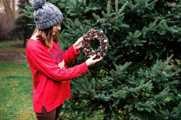 The pretty girl wearing cozy sweater with ornament of deer is hanging up colorful handmade Christmas wreath with cones and glistening balls on the tree outside