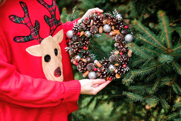 The pretty girl wearing cozy sweater with ornament of deer is hanging up colorful handmade Christmas wreath with cones and glistening balls on the tree outside