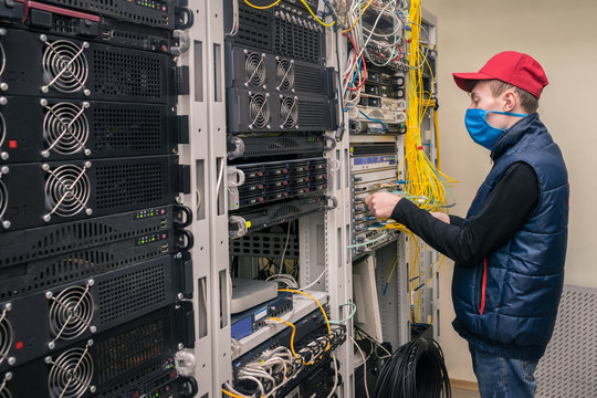 Young Technician In Medical Mask Switches Fiber Optic Wires On The Main Router. The Installer Works In The Server Room During Quarantine. Data Center Service.