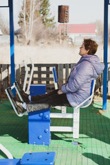 Middle-aged woman in outerwear does gymnastic exercises on free street exercise machines. The concept of a healthy lifestyle, fitness in quarantine and coronovirus