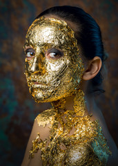 Girl with a mask on her face made of gold leaf. Gloomy studio portrait of a brunette on an abstract background.