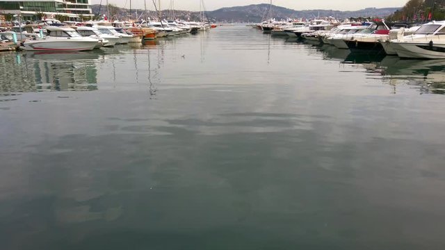 Cloudy foggy day. Private yachts and sailing boats with lowered sails standing on pier in bay. Reflections in water. People walk along promenade. Turkey, Istanbul, Tarabya, March 2020.
