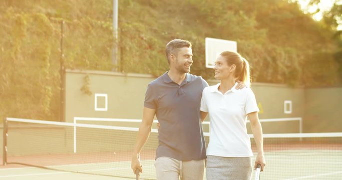 Caucasian happy attractive couple walking away from sport court after playing tennis in sunlight of sunset. Pretty woman and handsome man hugging and leaving pitch after game.