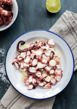 Overhead view of a delicious dish of cut cooked octopus over a potato bed. A part of another dish with some octopus arms are in the up left. A blurred image complete the scene.