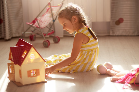 Close Up Portrait Of Beautiful Toddler Girl With Pigtails Sitting On Floor Playing With Toy Doll House, Toy Baby Carriage Near Window On Background., Female Kid Wearing Yellow Dress. Childhood Concept