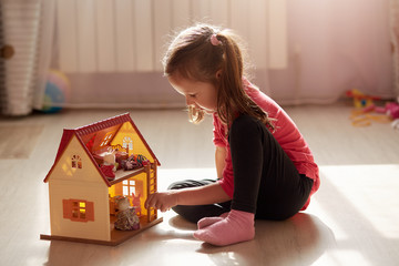 Indoor shot of liitle charming girl sits on floor with her doll house, wearing casual clothing, has...