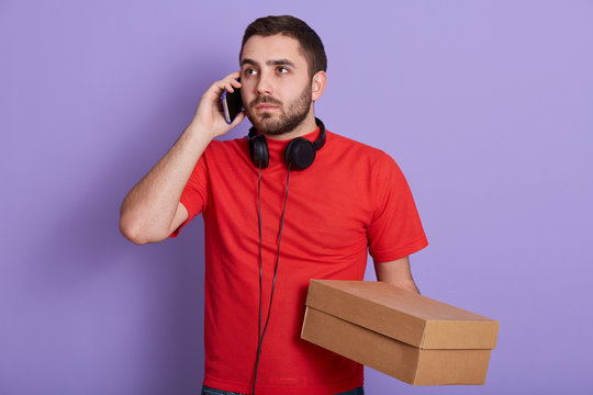 Portrait of hendsome delivery man wearing red t shirt talking on mobile phone while holding carton box in hands isolated over lilac studio background, looking at distance, has headphones around neck.