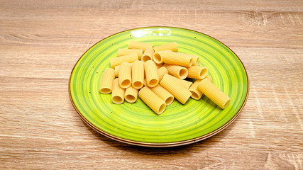 Uncooked raw rigatoni pasta on a green plate on wood background. Top view, vertical photo, selectiv focus, with copy space, space for text.