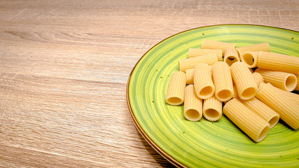 Uncooked raw rigatoni pasta on a green plate on wood background. Top view close-up, vertical photo, selectiv focus, with copy space, space for text.