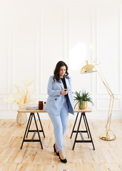 A serious business working freelance girl a woman with black hair in a pale blue suit working on a laptop and phone at a wooden Desk against a white wall in the home office