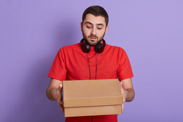 Delivery Concept. Portrait of curious caucasian handsome bearded delivery man wearing red casual t shirt, holding carton box package, isolated over lilac studio background. Copy space for advertisment