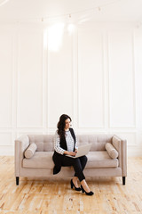 Serious business working freelance woman with black hair in a business suit working on a laptop and phone on a gray sofa against a white wall in the house, business portrait