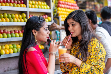 Young happy smiling Indian girls drinking or holding glass of orange juice outdoor in the market with colorful background of fruits. Urban healthy lifestyle.