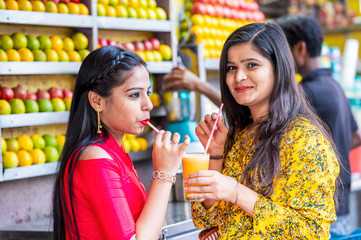 Young happy smiling Indian girls drinking or holding glass of orange juice outdoor in the market with colorful background of fruits. Urban healthy lifestyle.