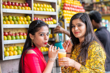 Young happy smiling Indian girls drinking or holding glass of orange juice outdoor in the market with colorful background of fruits. Urban healthy lifestyle.