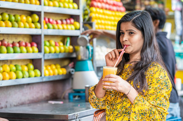 Young happy smiling Indian woman drinking orange juice outdoor in the market with colorful background of fruits.