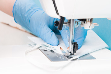 a woman in blue disposable medical gloves sews a protective mask for the face from cotton gray fabric on a sewing machine, blue background, the process of sewing masks, focus on fabric
