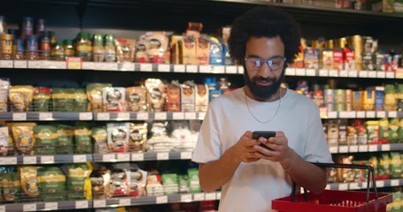 Handsome bearded man in glasses using smartphone while doing shopping with basket. Mature guy in 30s checking list on mobilephone while standing in food store section.