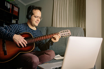 Boy doing an online course to learn how to play the classical guitar. He is sitting on the sofa at...