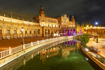 Renaissance building in Plaza de Espana in Seville, Andalusia, Spain, reflects on channel of Guadalquivir river. Scenic Spain Square illuminated at night, popular landmark.