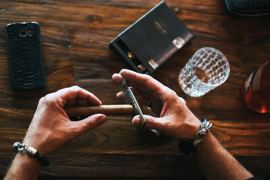 Men's Hands Cut A Cigar With Scissors. Wooden Table.