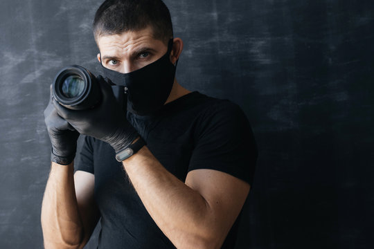 Photographer in a black mask, a T-shirt and gloves, holds the camera in his hands on a dark background. Concept Photo business and coronovirus.