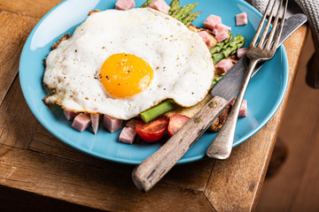 Healthy breakfast or lunch with fried egg, bread toast, green asparagus and tomatoes on blue plate on old wooden table