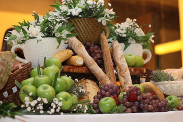 Mesa de comidas, com maças, pães, uvas e decoração de flores brancas.