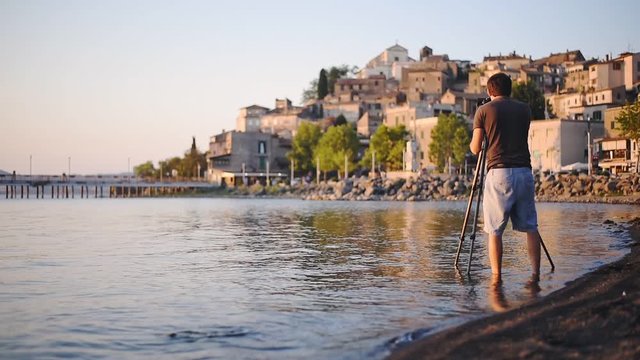 A Man Wearing Shorts Standing In The Lake Bracciano Shore With His Camera Capturing The Beauty Of Castello Orsini-Odescalchi In Rome, Italy. -wide shot