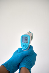 Human hand with blue disposable glove hold a digital infrared thermometer for body temperature scan...