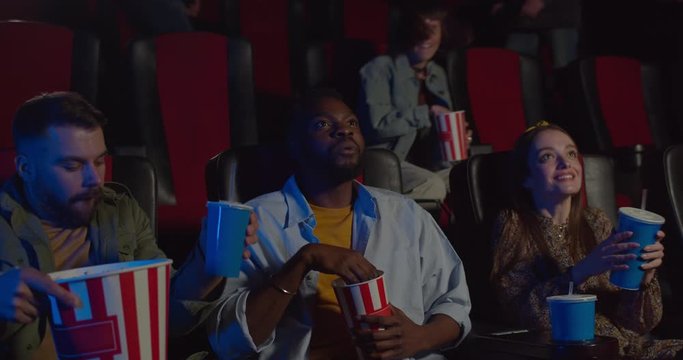 Excited spactators starting hold seats while film already started. Young african eating popcorn and watching film while other people taking their seats. Concept of leisure and entertainment.