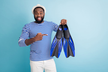 Photo of funny dark skin guy hold diver flippers excited to start sinking direct finger best mark model fins wear white sun cap striped sailor shirt shorts isolated blue color background