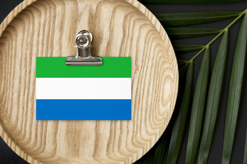 Sierra Leone flag tagged on wooden plate. Tropical palm leaves monstera on background. Minimal national concept.