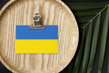 Ukraine flag tagged on wooden plate. Tropical palm leaves monstera on background. Minimal national concept.