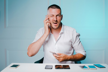 Young winking man holding two phones near his ear. The collection of mobile phones in front of him.