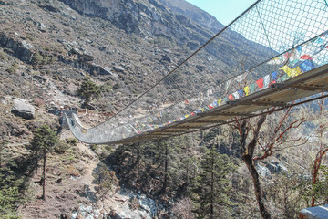 One of many pedestrian suspension bridges with waving prayer flags in Himalayas on the way to Everest Base Camp in Nepal. Clear blue sky. Side view. Theme of trekking in Himalayan mountains. - obrazy, fototapety, plakaty