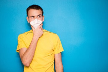 Young handsome man in medical mask and yellow t-shirt think how to dispose of problem isolated on blue background. Makes up an idea, solution of problem.
