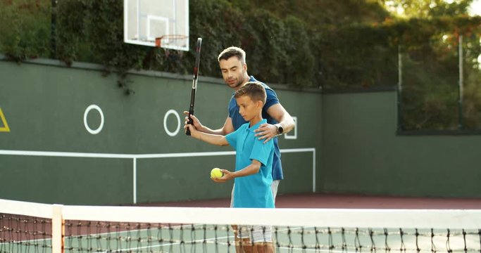 Caucasian handsome happy dad teaching his little cute teenage son to play tennis at sport pitch on sunny summer day. Kid learning to hit ball with racket from male instructor outdoor.