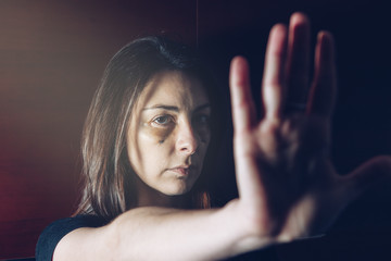 Abused Caucasian woman with bruises and stitches on her face, with her arm outstretched her hand gestures to stop. Soft light coming in. Concept of domestic violence victims