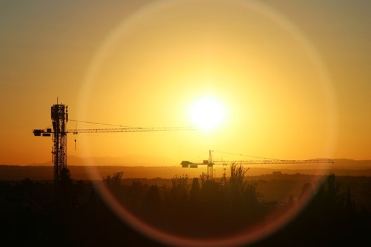 Torres gr&uacute;a de construcci&oacute;n al atardecer, con el sol poni&eacute;ndose al fondo generando un bonito halo.