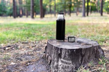 Black thermos bottle and a cup on a stump in a park in early fall