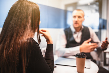 Rearview of businesswoman and businessman having a conversation during a coffee break in a cozy coffee-point in a modern office, selective focus on the woman, laptop and disponsable cup on the table